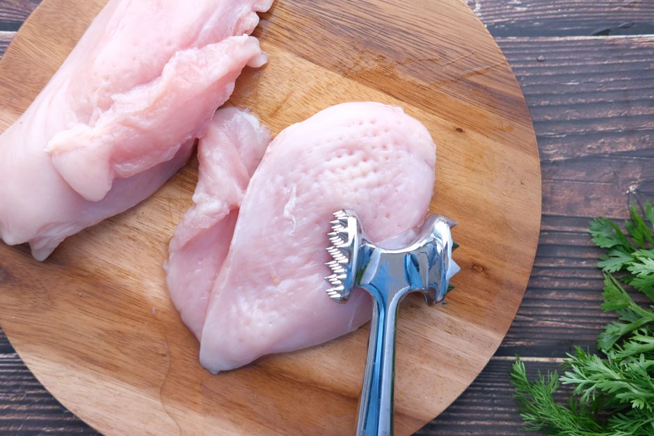 Raw chicken breast on a wooden cutting board with a meat mallet
