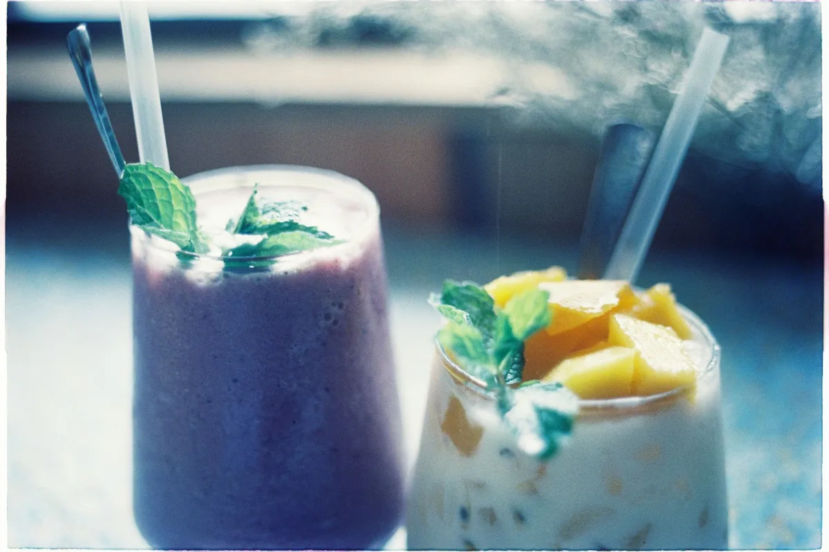 A blueberry smoothie and a fruit-topped drink in glasses with straws