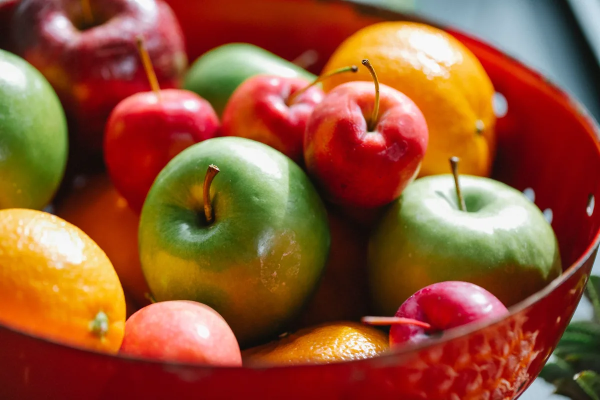A bowl of whole red apples and oranges on a bright wooden table