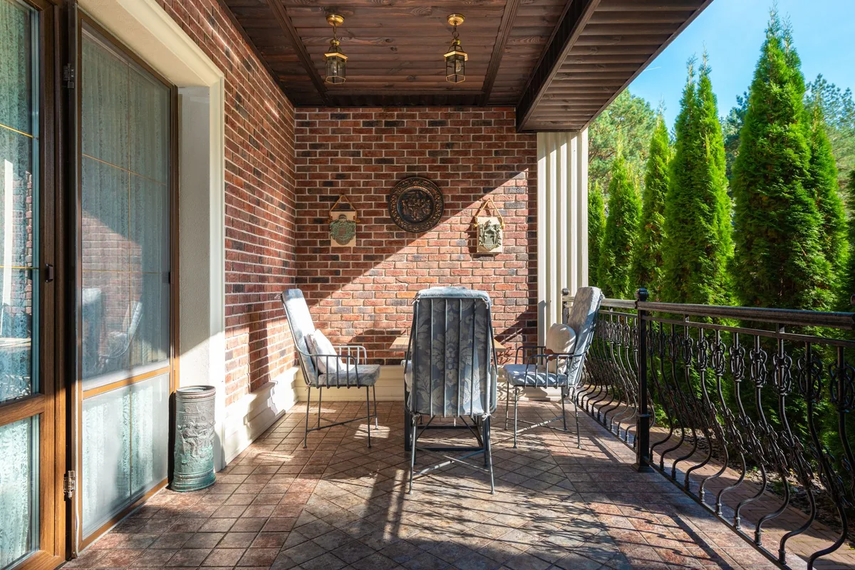 A covered balcony with chairs and a table beside a brick wall and railing