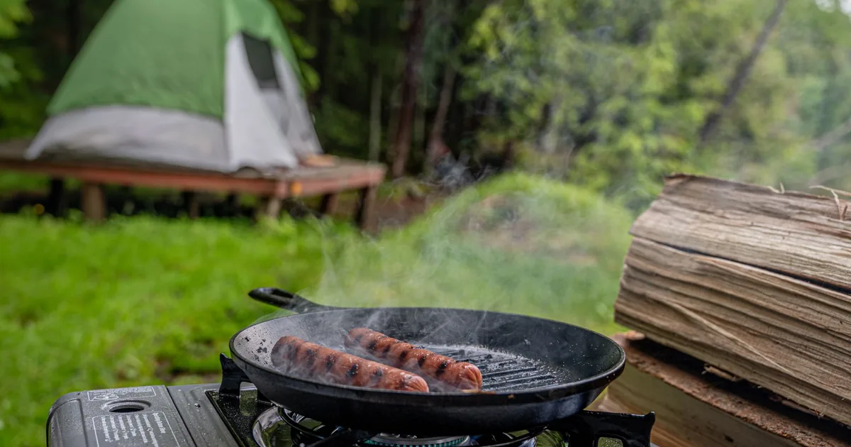A camp stove with sausages in a pan beside a green tent on a grassy site