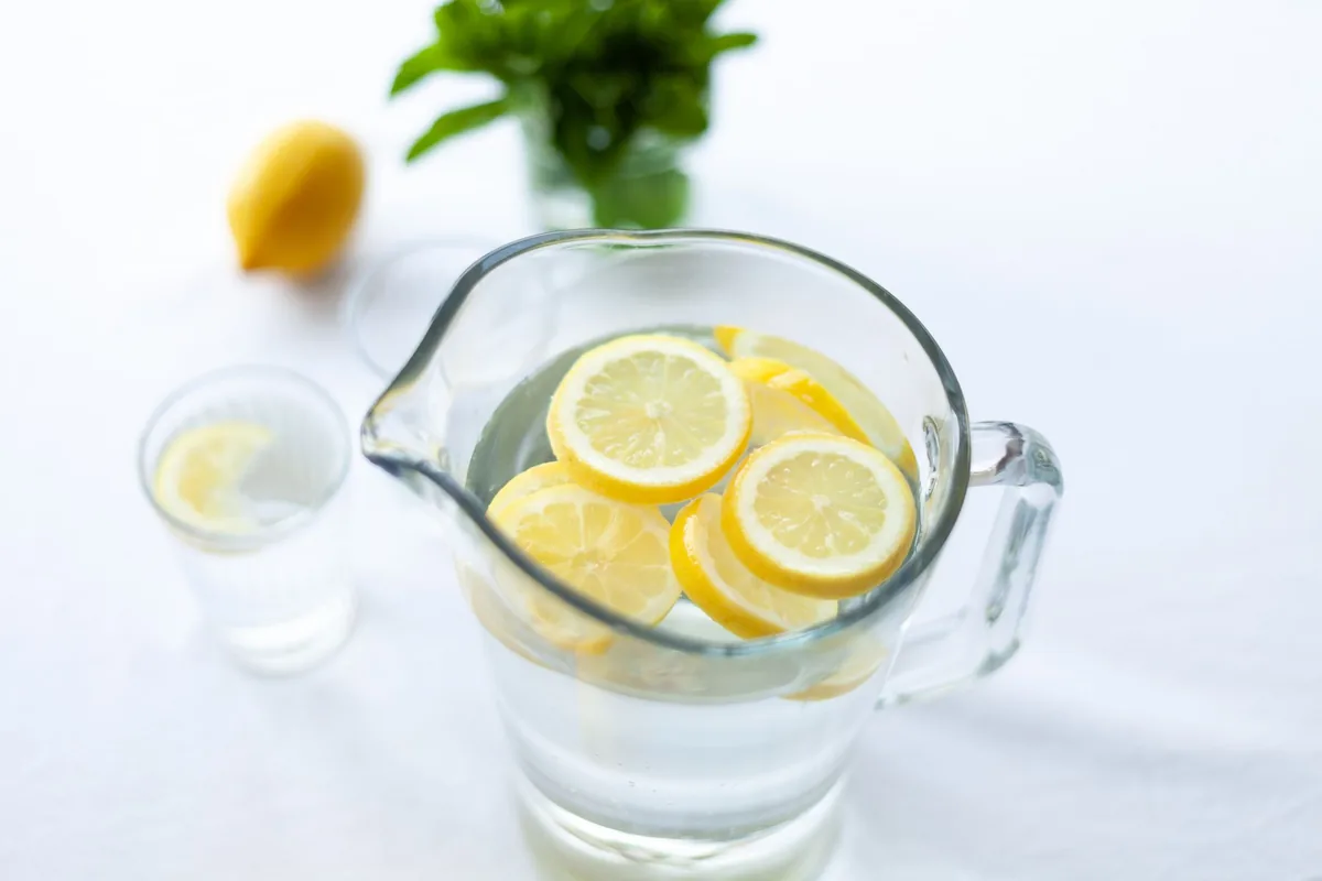A clear glass pitcher of lemonade with lemon slices on a sunny table