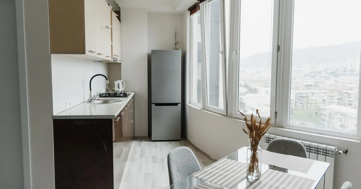 A compact white refrigerator standing in a bright, tidy apartment kitchen corner