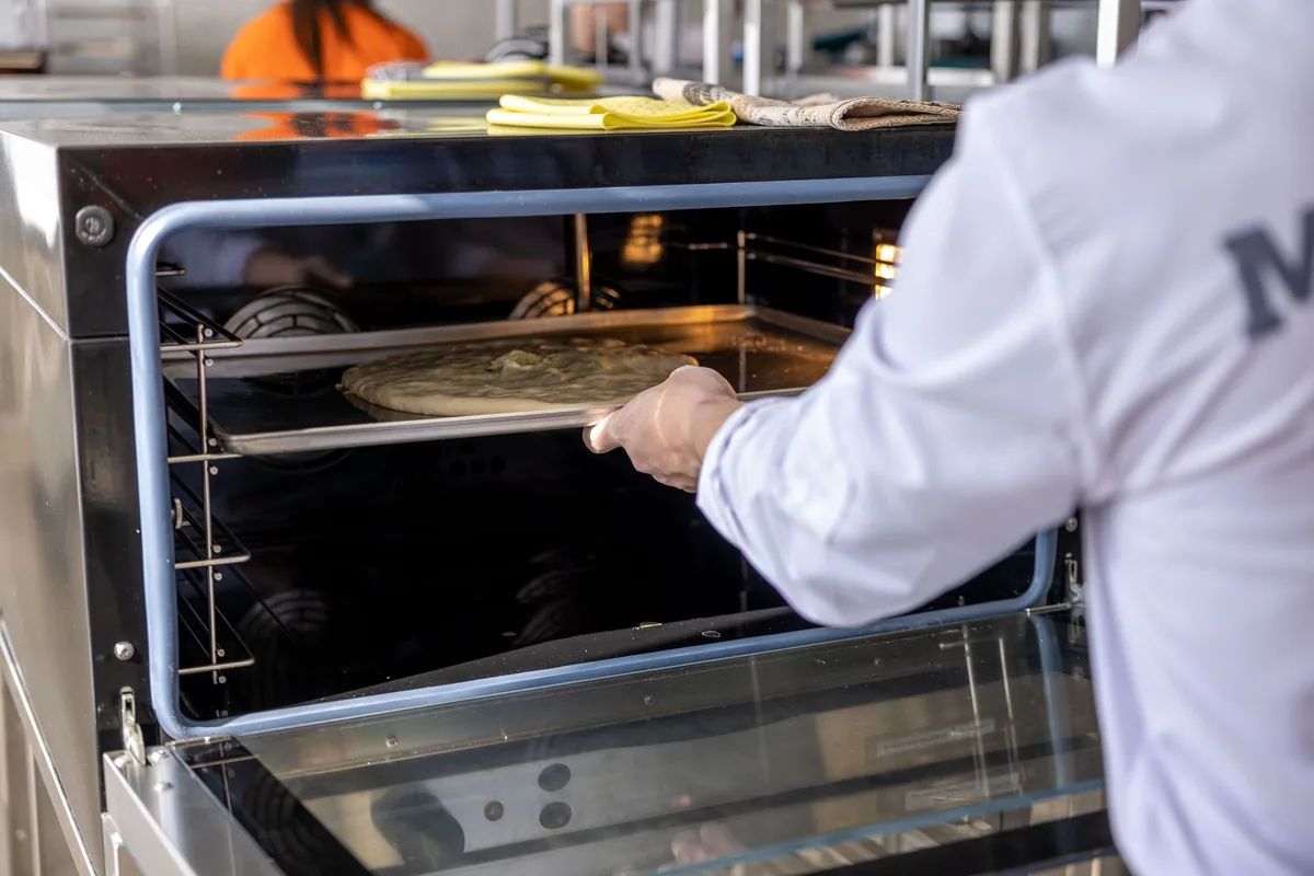 A cook wearing a blue oven glove sliding a tray into a commercial oven