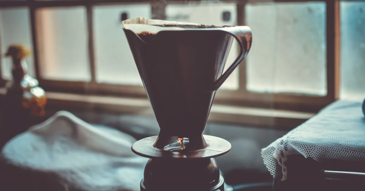 A metal pour over dripper on a stand by a window