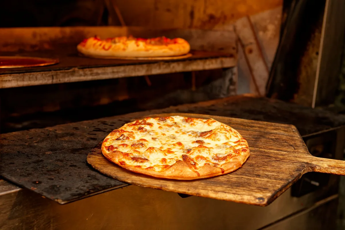 Pizzas baking in a deck oven, one on a wooden peel