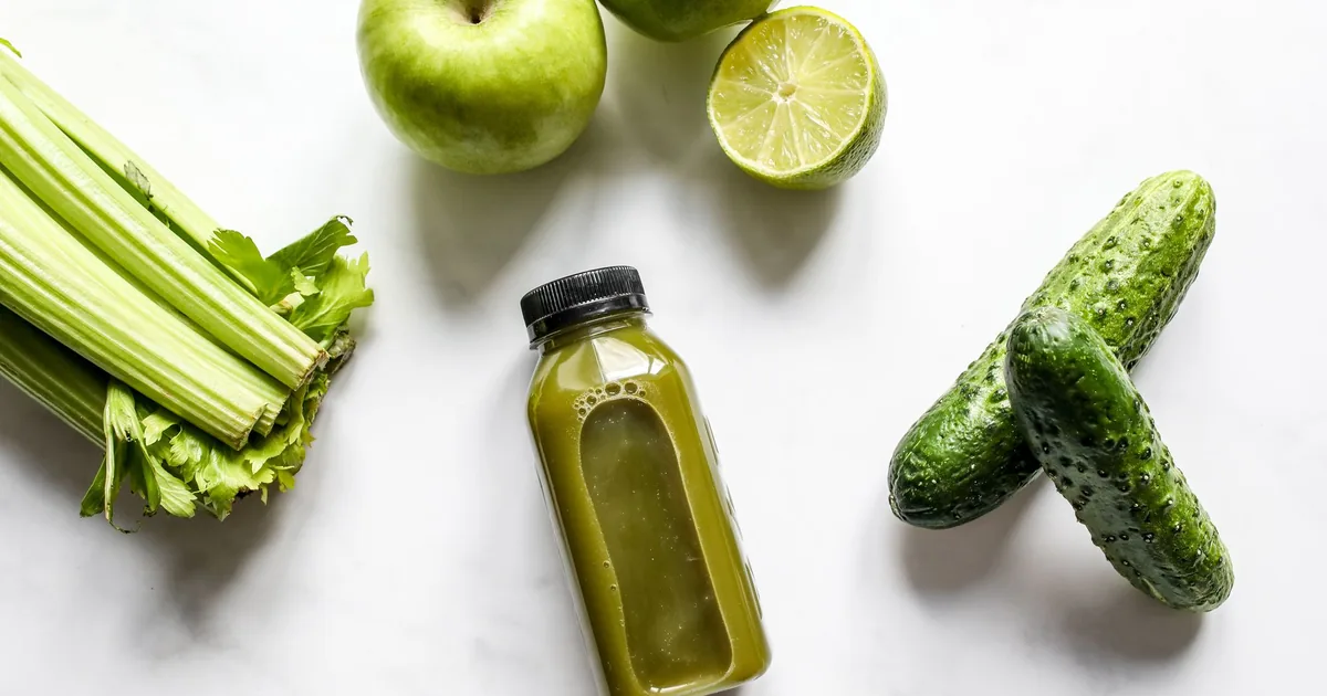 A glass of bright green juice on a light kitchen counter beside whole green apples and celery stalks