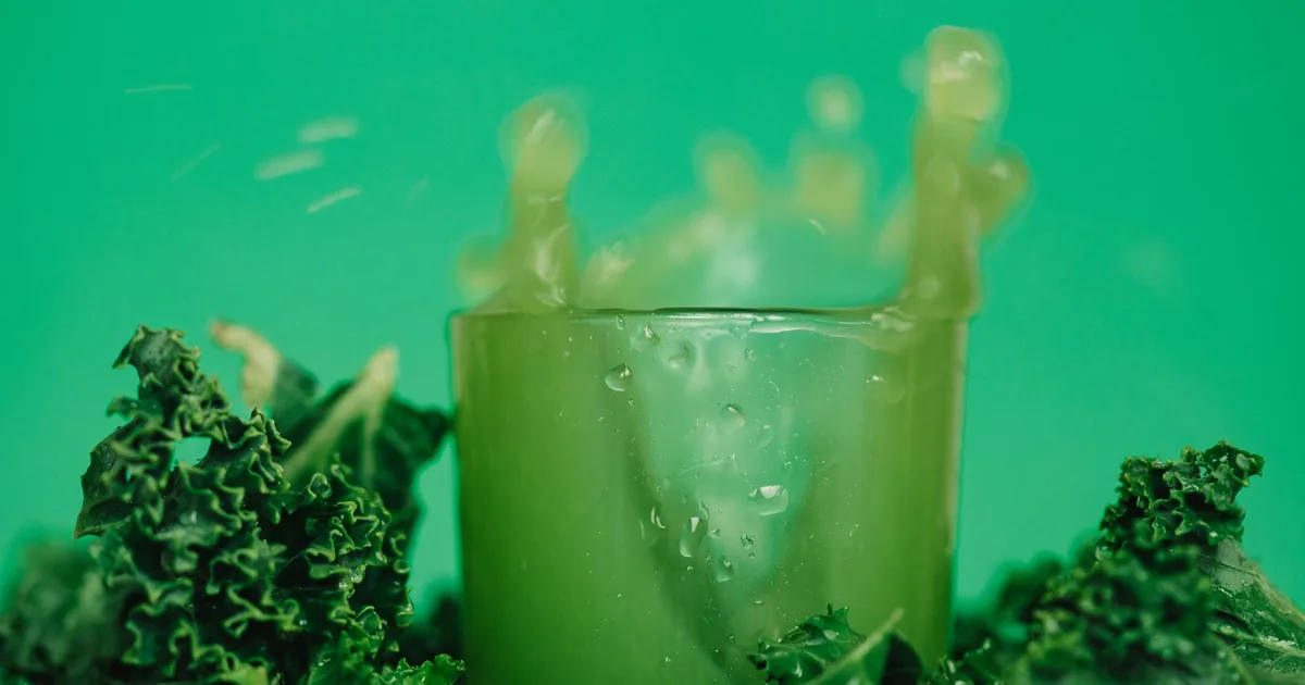 A glass of bright green juice on a wooden board surrounded by fresh kale, spinach and celery