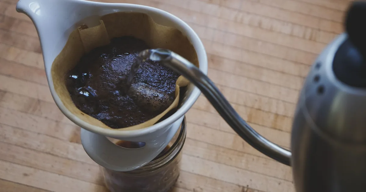 A gooseneck kettle beside a pour-over coffee dripper and glass carafe on a light wooden counter