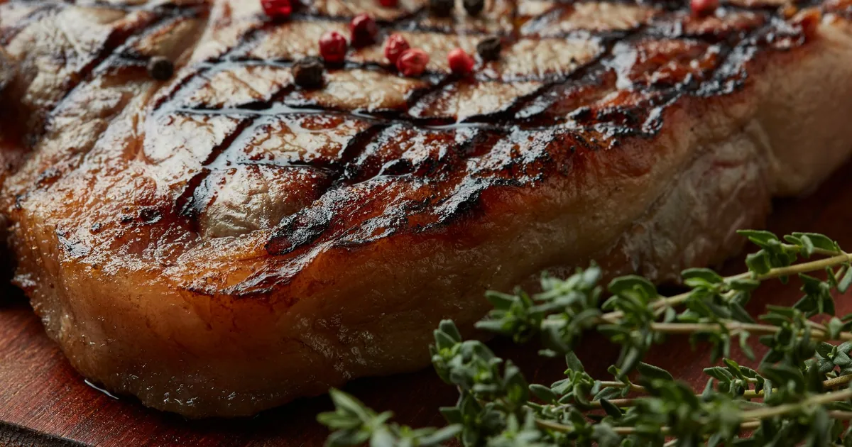 A grilled steak with dark grill marks resting on a wooden board beside grilled vegetables