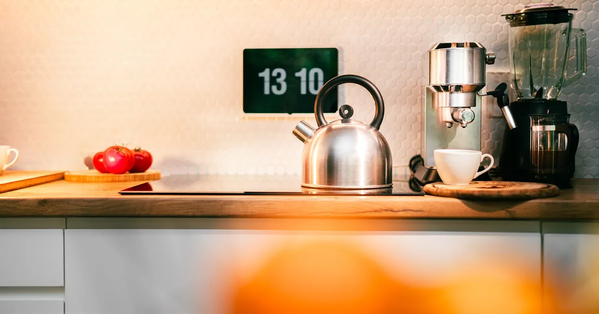 A kitchen counter with a blender, a kettle and a digital clock