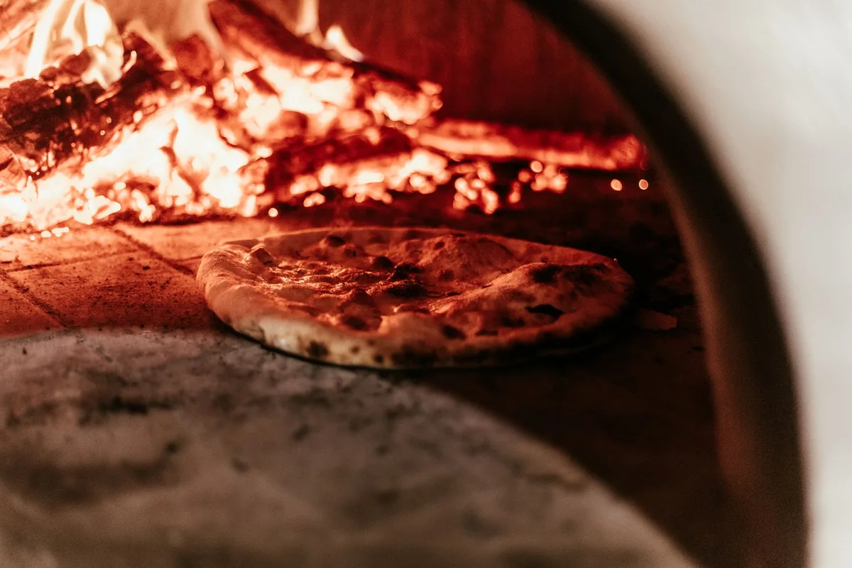 A pizza baking beside glowing embers in a wood-fired oven