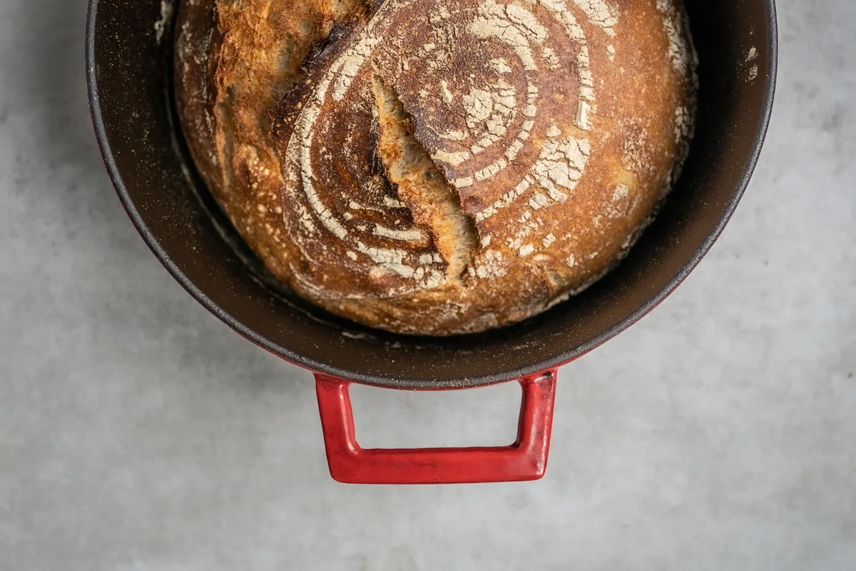 A round crusty loaf of bread resting beside a lidded baking pot