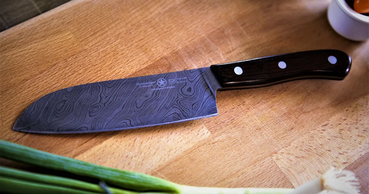 A santoku-style kitchen knife resting on a wooden cutting board beside neatly chopped vegetables
