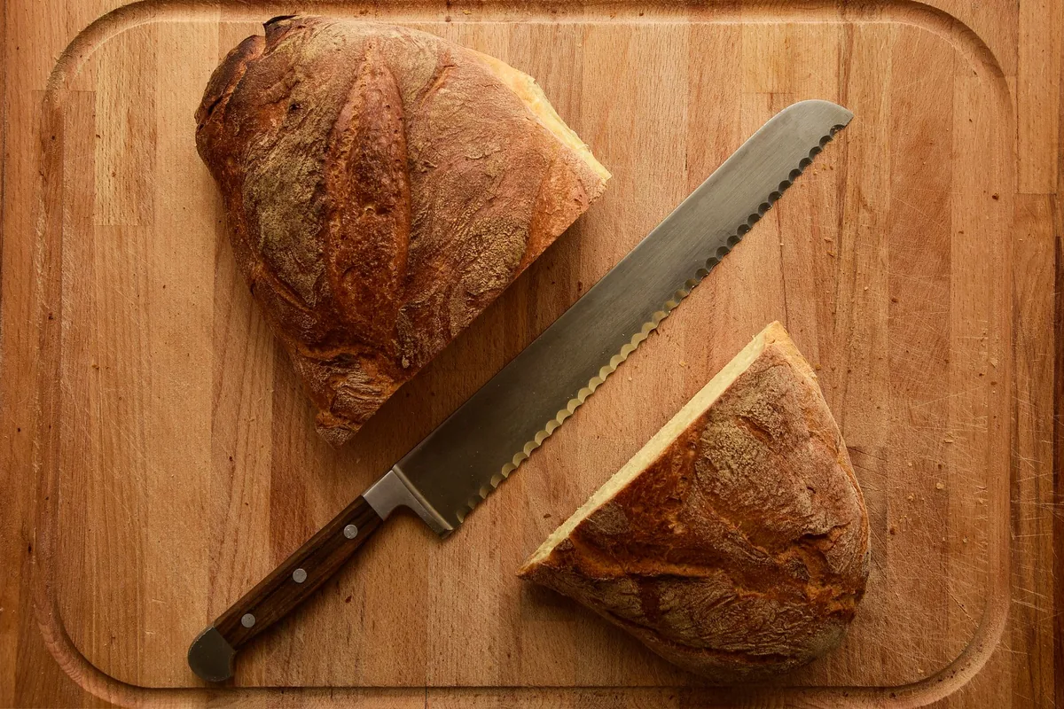 A serrated bread knife beside a crusty loaf on a wooden board