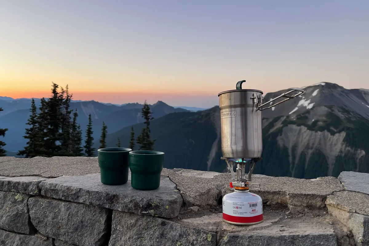 A small gas canister stove on a stone wall overlooking mountains at dusk
