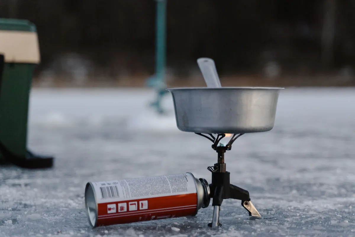 A single-burner camp stove on a gas canister holding a metal pot, standing on bare ground