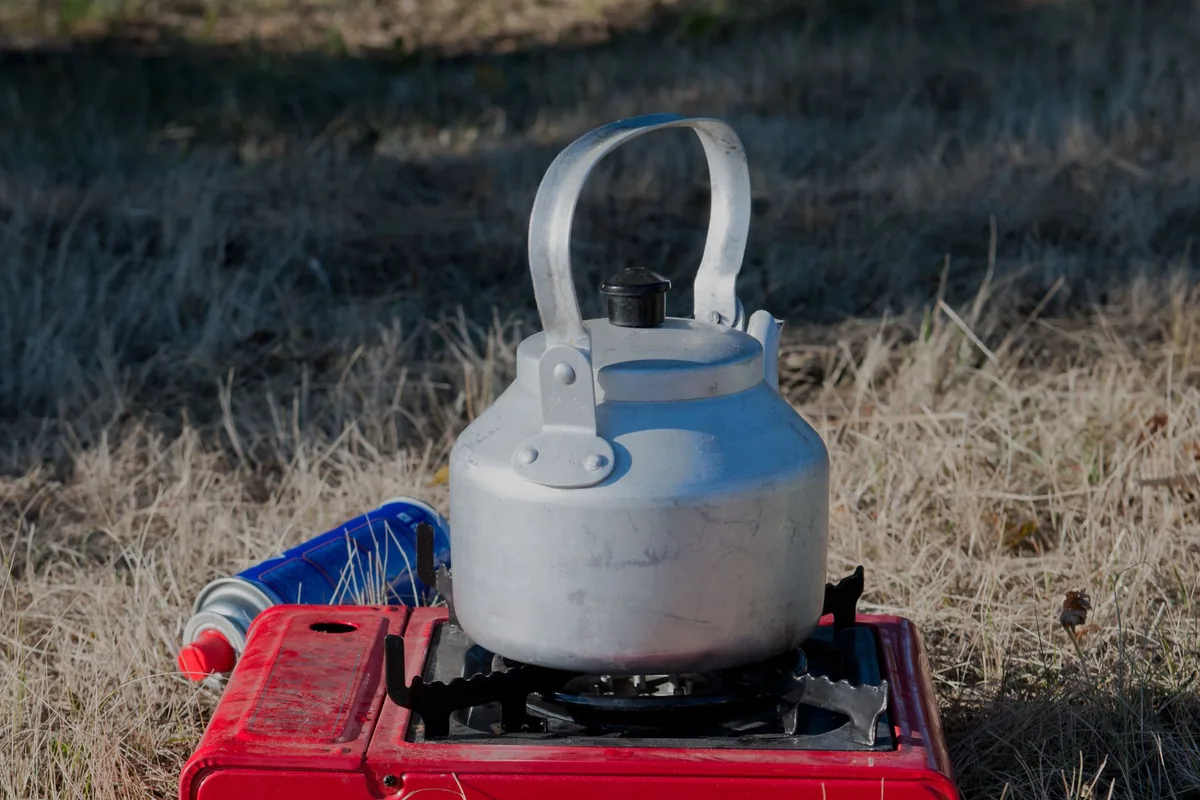 A small red portable gas stove with a kettle on dry grass outdoors