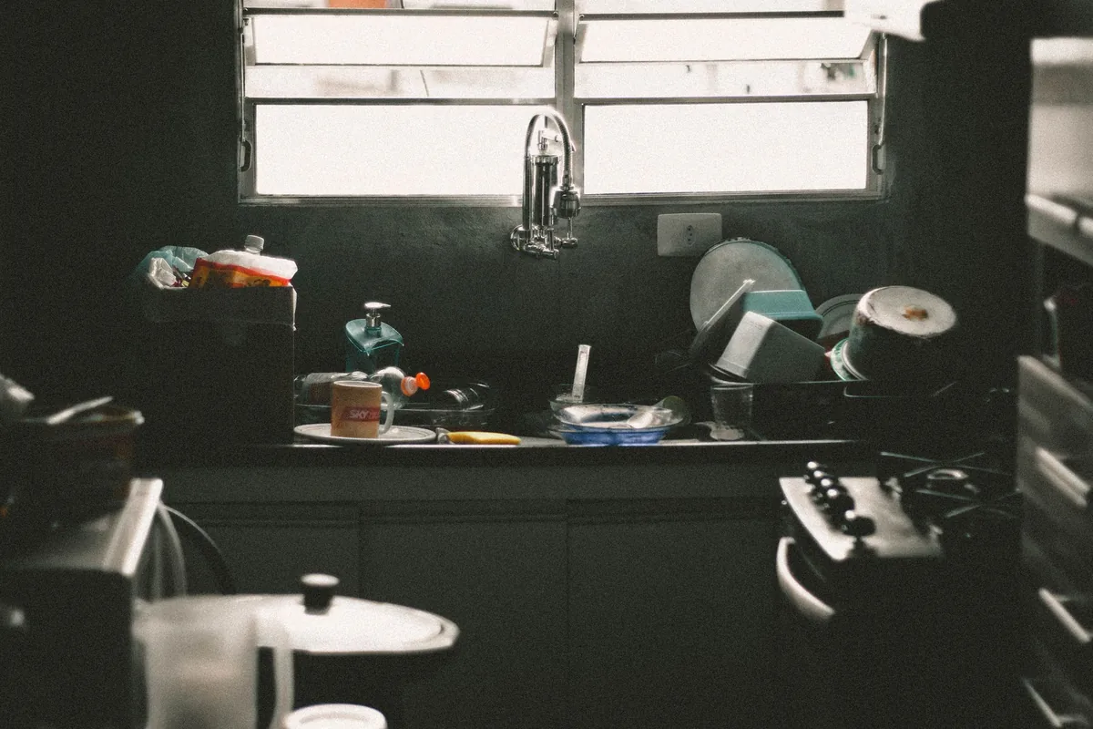 A sink piled with used dishes below a window in a dim kitchen