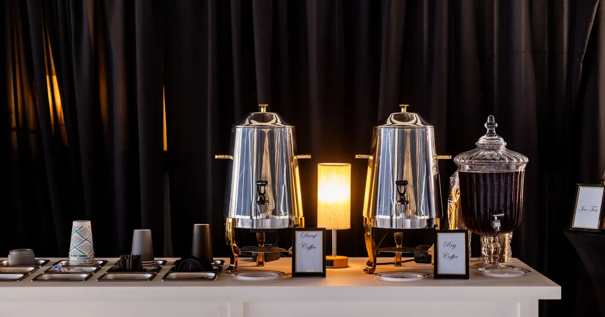 A stainless steel coffee urn on a catering table beside rows of white cups and saucers