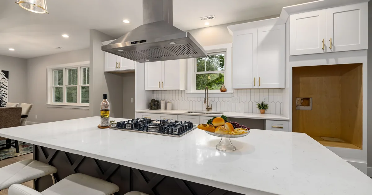 A stainless steel range hood mounted beneath white wall cabinets above a wide cooktop in a bright kitchen