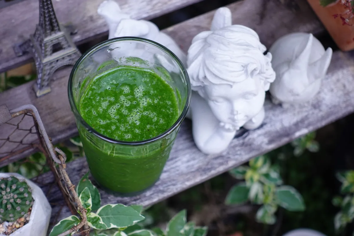 A tall glass of bright green smoothie on a white table with a few spinach leaves beside it