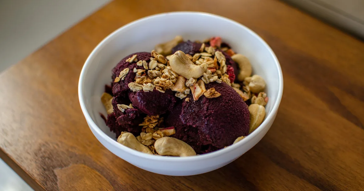 A thick purple acai bowl topped with sliced banana, berries and granola on a light table