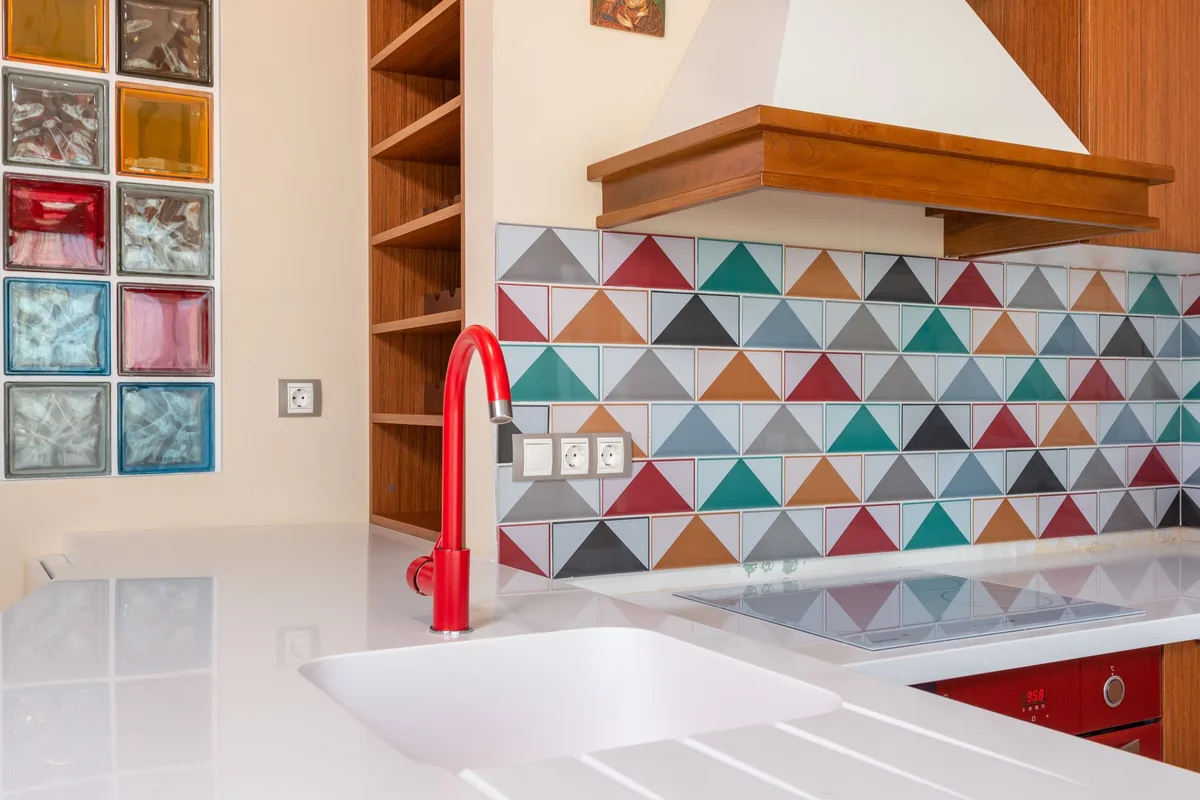 A white kitchen with a red faucet and a colourful tile backsplash