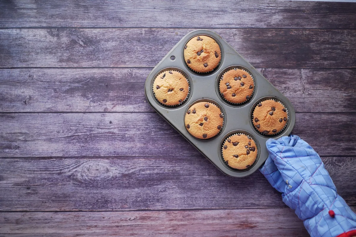 Freshly baked muffins in a six-cup tin beside a blue oven mitt