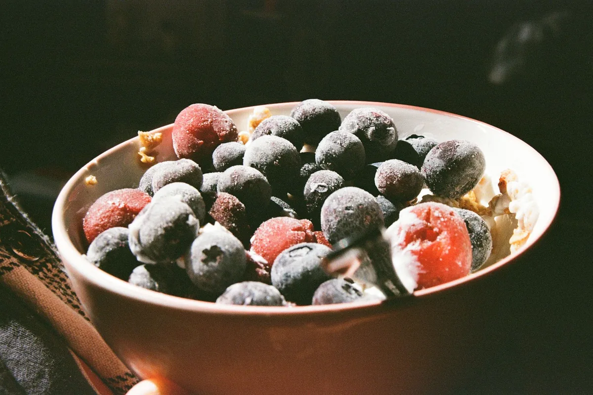 A white bowl heaped with frozen mixed berries dusted with frost on a light worktop