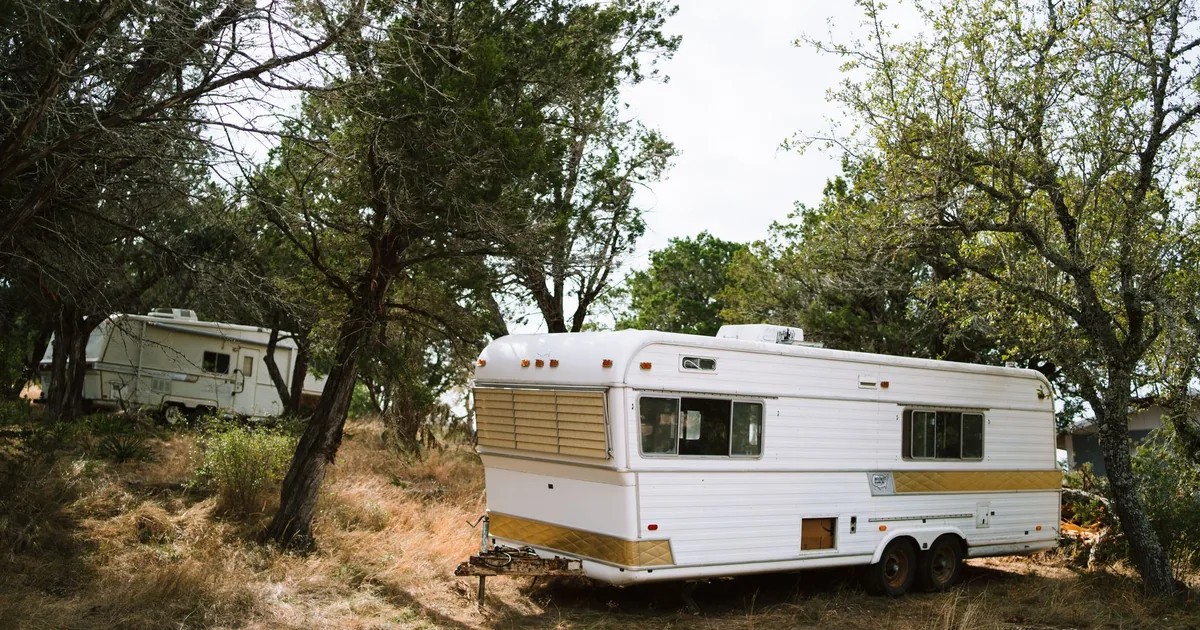 A white travel trailer parked on a gravel campsite surrounded by green trees on a sunny day