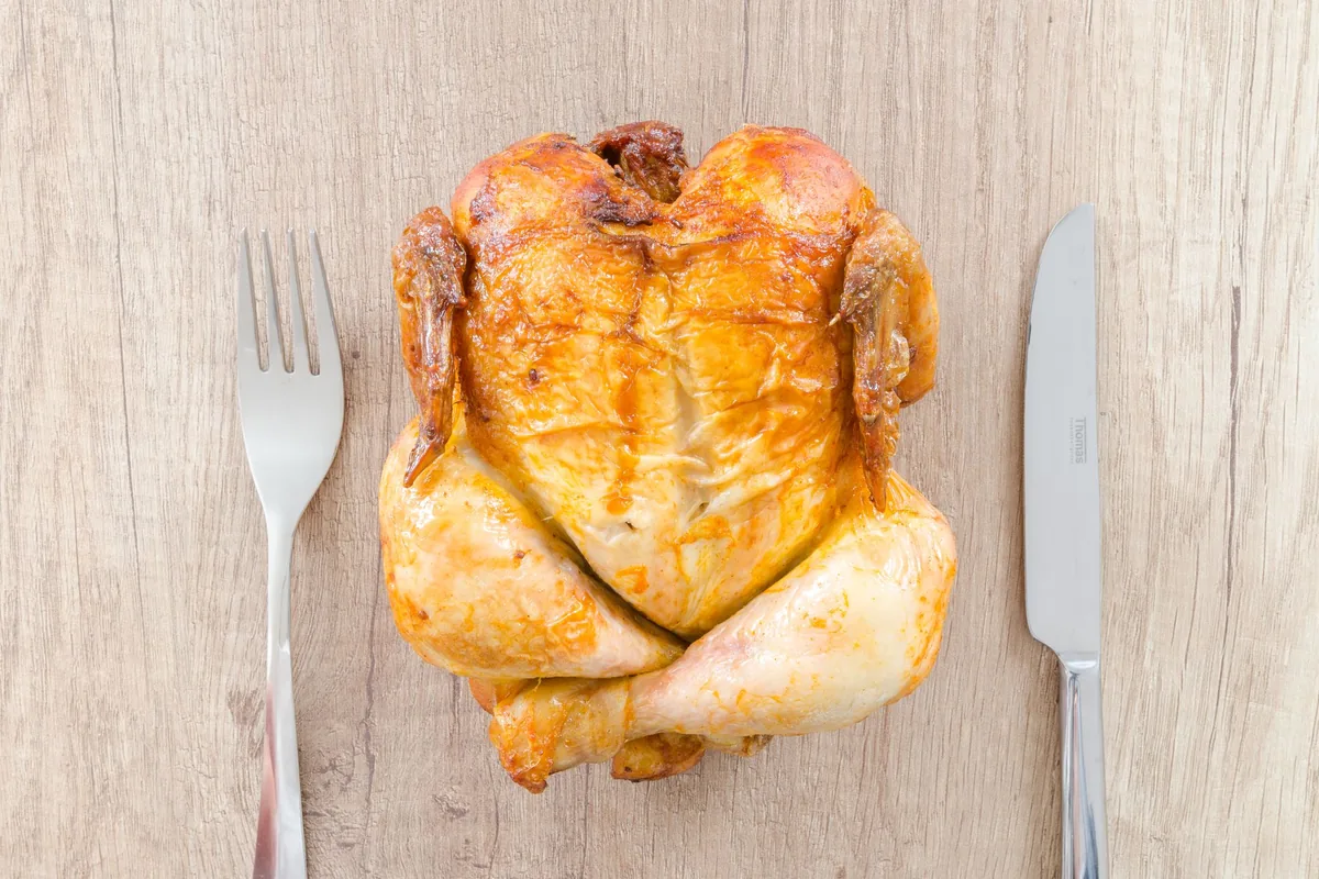A whole roast chicken on a wooden board between a fork and a knife