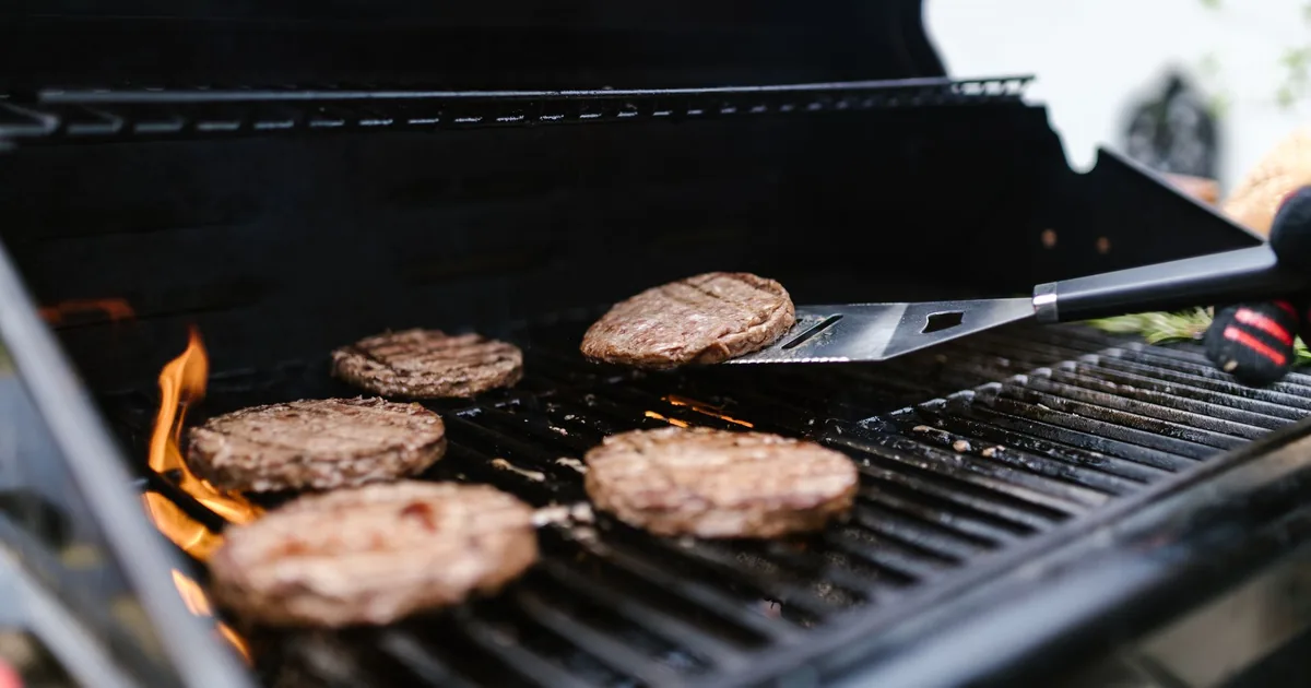 Burger patties and sliced peppers cooking on the grate of a small gas grill outdoors in daylight