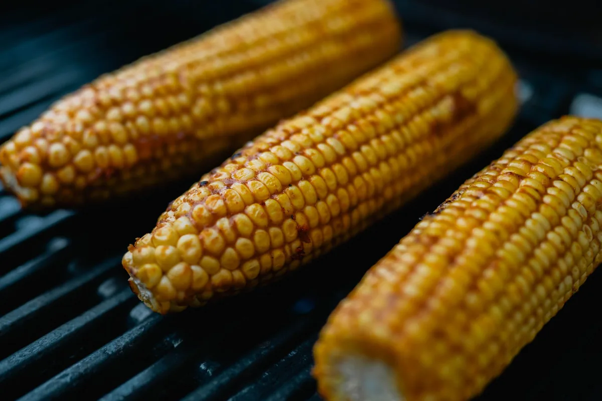 Corn cobs and sliced peppers charring on an outdoor grill grate in daylight