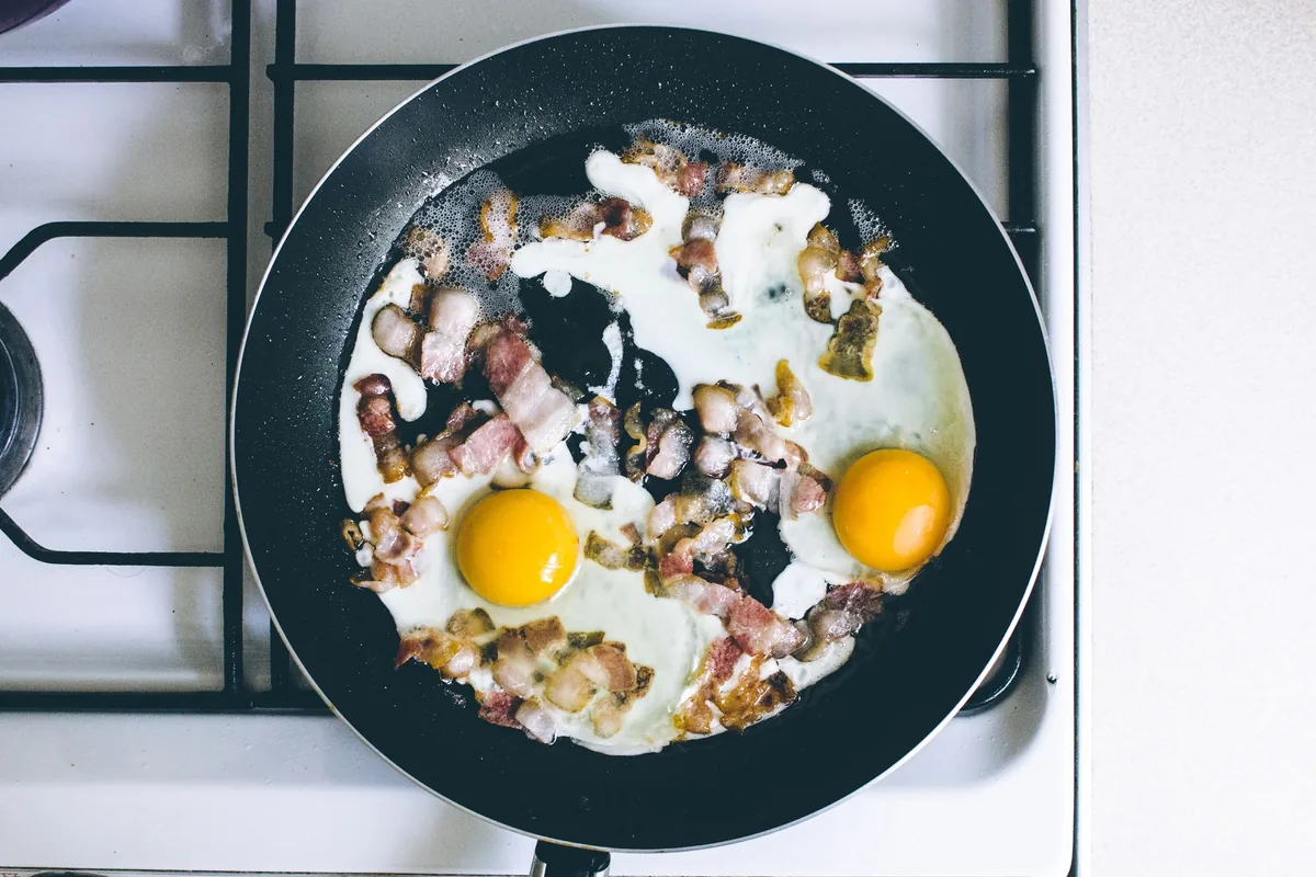 Fried eggs and bacon in a black frying pan on a white gas stove, seen from above