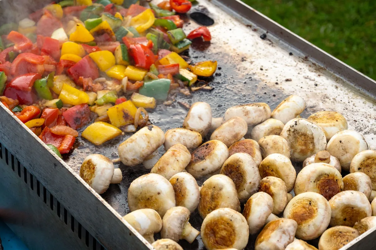 Peppers, mushrooms and onions searing on a wide outdoor flat-top griddle