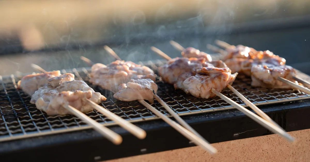 Rows of chicken skewers cooking over a small rectangular charcoal grill on an outdoor table in daylight