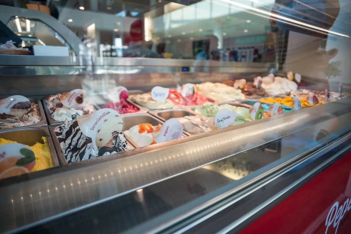 Rows of gelato tubs in a refrigerated display case with flat paddles in each