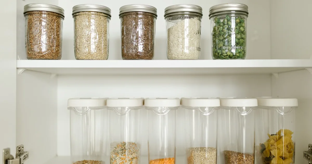 Rows of glass mason jars filled with rice, beans, pasta and oats on a bright pantry shelf