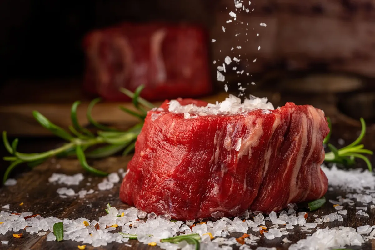 Salt falling onto a thick raw steak on a dark board