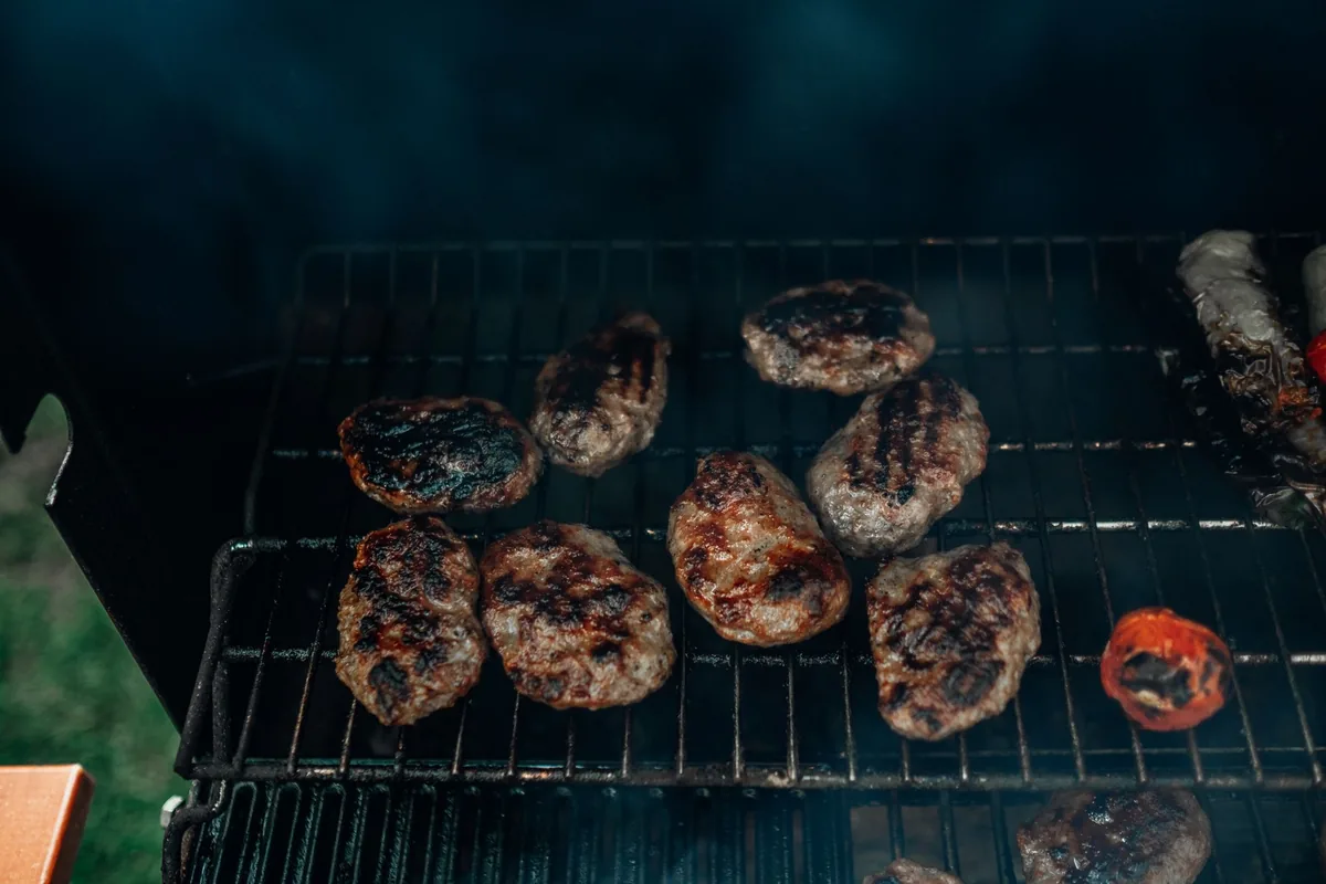 Several burger patties and sausages cooking on the round grate of a small charcoal grill