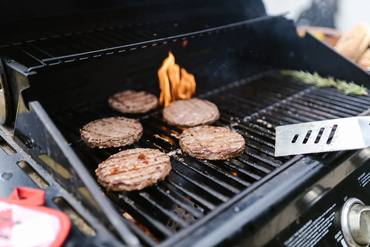 Several burger patties browning on the grates of a gas grill with the lid open