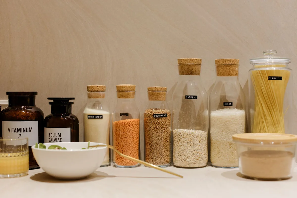 Several glass jars of dry pasta, lentils and grains lined up on a wooden counter