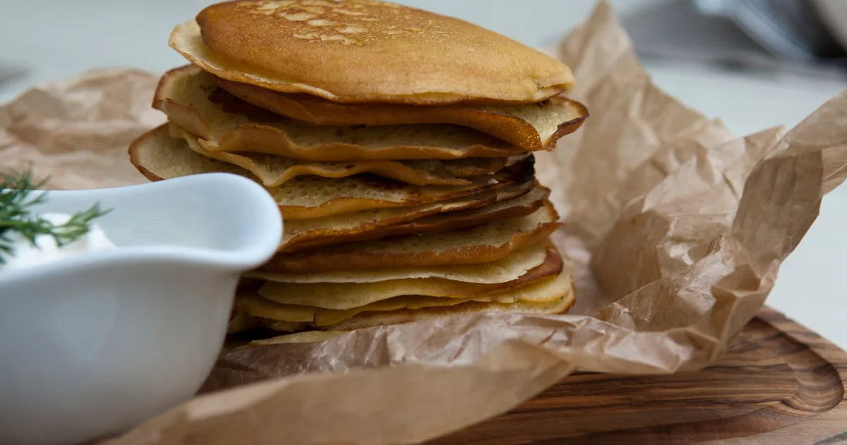 A tall stack of golden pancakes on baking paper beside a small white jug
