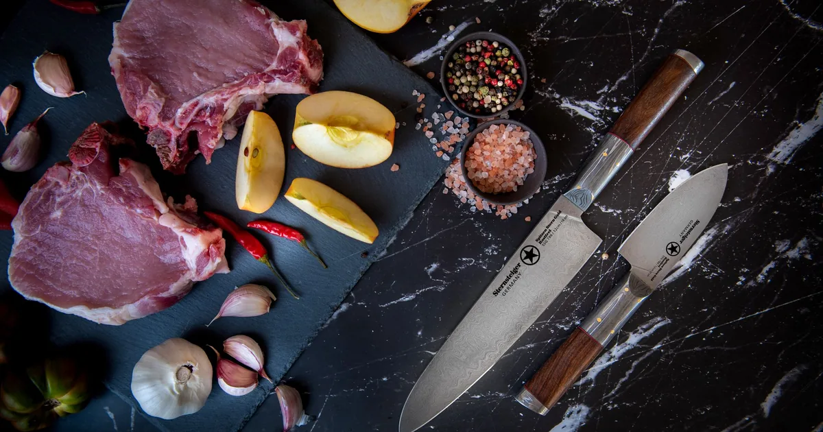 Two knives beside raw steak, garlic and spices on dark marble