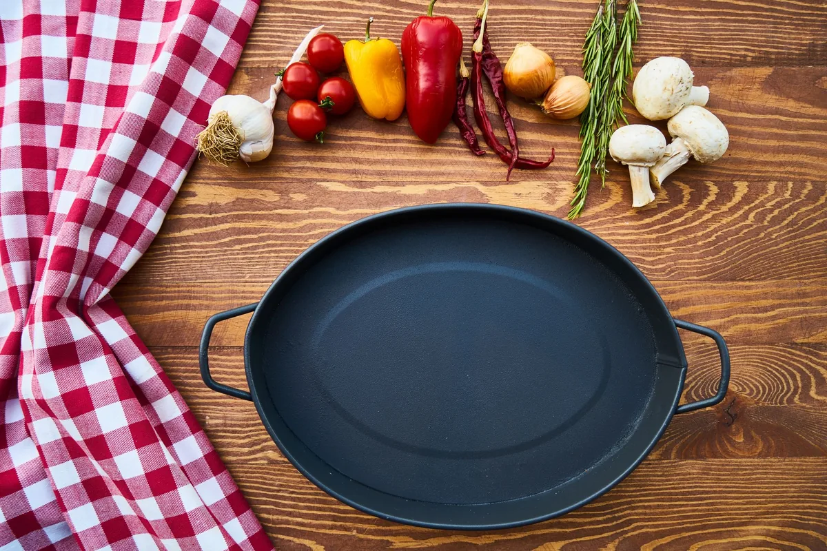 An oval black pot on a wooden table beside fresh vegetables