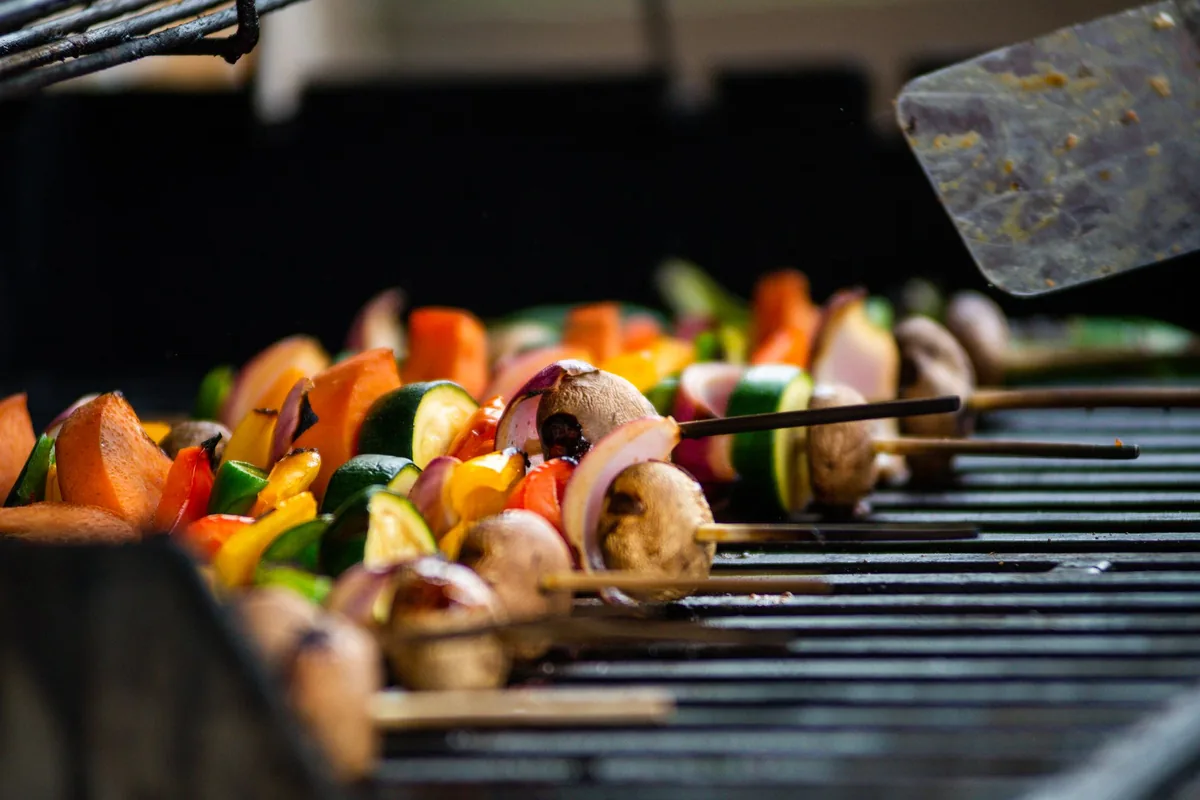 Skewers of peppers, onions and mushrooms browning on a metal grill grate in bright daylight