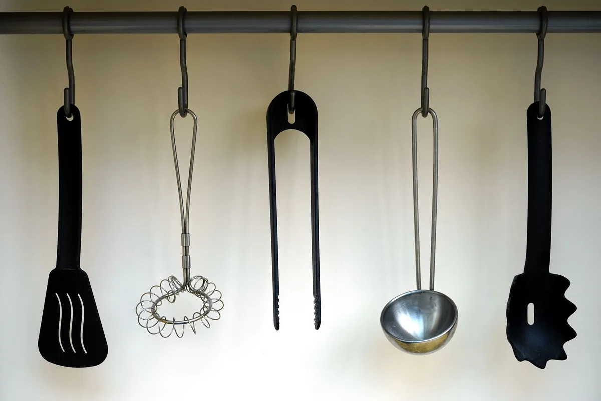 Stainless kitchen utensils hanging from metal S-hooks on a rail