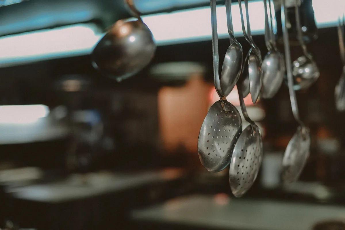 Steel kitchen utensils including whisks hanging from a rail on a light tiled wall