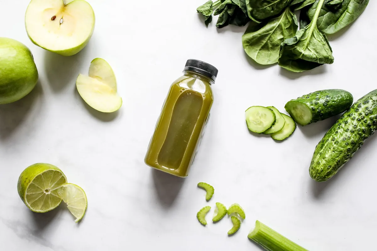 Two clear glass bottles of green vegetable juice on a bright worktop beside spinach leaves